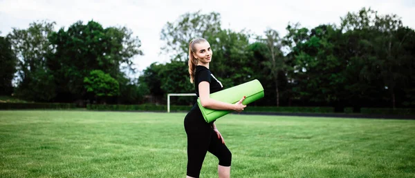 健康漂亮的女人在夏季公园慢跑后喝水 在运动后出汗 训练后的运动员 小女孩手里拿着一块运动垫 这个女孩给全班同学表演 — 图库照片
