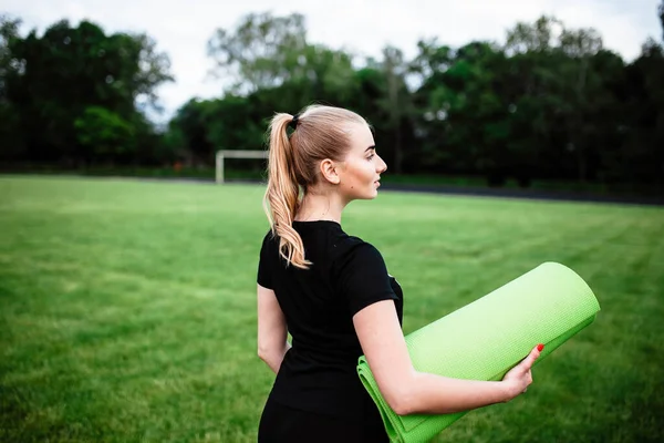 健康漂亮的女人在夏季公园慢跑后喝水 在运动后出汗 训练后的运动员 小女孩手里拿着一块运动垫 这个女孩给全班同学表演 — 图库照片