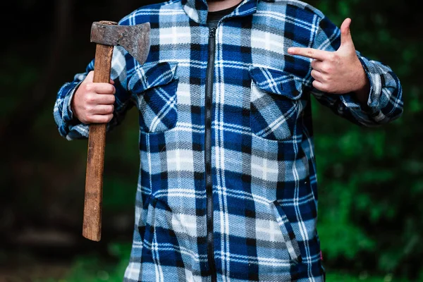 A lumberjack works in the forest. Lumberjack with an ax in the summer forest. The lumberjack is holding an ax. The lumberjack holds an ax on his back. The lumberjack shows the class. Ax in hand.