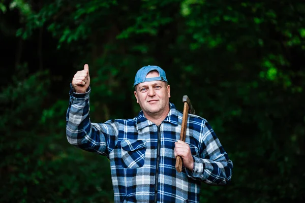 A lumberjack works in the forest. Lumberjack with an ax in the summer forest. The lumberjack is holding an ax. The lumberjack shows the class. Ax in hand.The lumberjack holds an ax on his back.Forest.