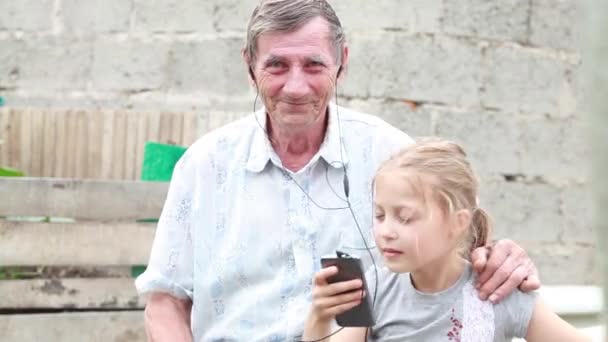 Abuelo y nieta escuchando música en auriculares y bailando sosteniendo un teléfono celular en el parque — Vídeo de stock