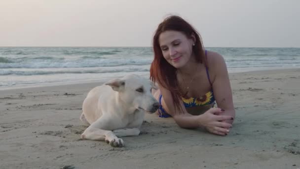 Una joven se acuesta con un perro blanco en la playa. 4K — Vídeo de stock