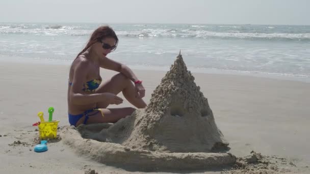 Hermosa chica con un castillo de arena incorporado en una playa tropical, mujer en el fondo de un mar de verano castillo de arena. 4K — Vídeos de Stock