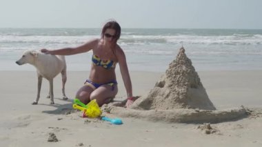 Bir kız bir mayo beyaz bir köpek ile kumsalda kumdan kale oluşturur. 4k