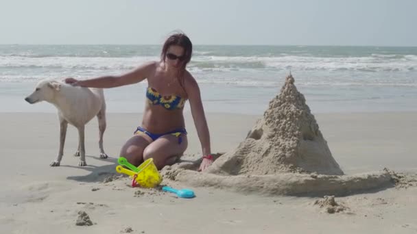 En flicka i en baddräkt med en vit hund bygger ett sandslott på stranden. 4k — Stockvideo