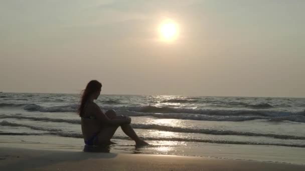 La chica está sentada al atardecer en la playa soñando. 4K — Vídeos de Stock