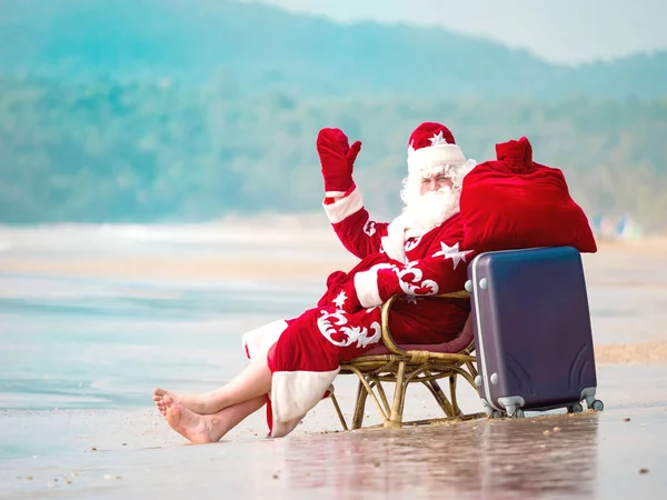 Santa con una maleta apoyada en el océano levantando la mano — Foto de Stock