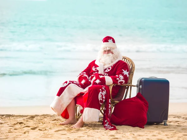 Święty Mikołaj siedzi na krześle boso z wielką świąteczną torbą pełną prezentów i walizką na piaszczystej plaży oceanu. — Zdjęcie stockowe