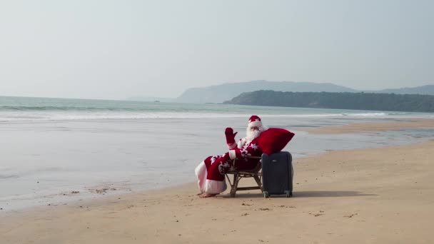 Santa Claus with a suitcase and a red bag sits in a chair by the ocean and waves his hand. — Stockvideo