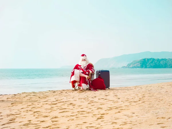 Noel Baba bir bavul ve hediye dolu bir çantayla sahilde oturuyor. — Stok fotoğraf