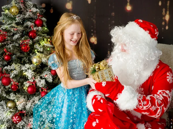Noel Baba ve mavi elbiseli kız yeni yıl ağacının yanında bir hediye. — Stok fotoğraf