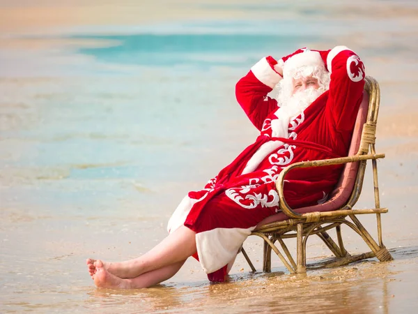 Santa Claus está sentado en un sillón con las manos detrás de la cabeza y descansando en la orilla del mar . — Foto de Stock