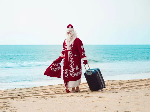 身穿红色衣服，提着手提箱和礼物袋的圣诞老人沿着海滨走着. — 图库照片