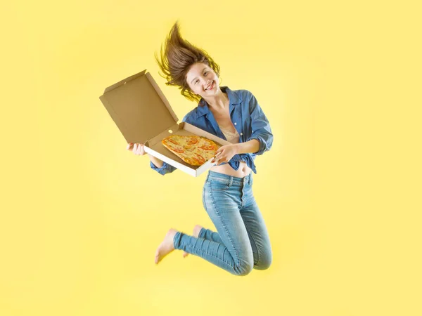 Hermosa joven en jeans sonriendo sostiene una caja de pizza sobre un fondo amarillo — Foto de Stock