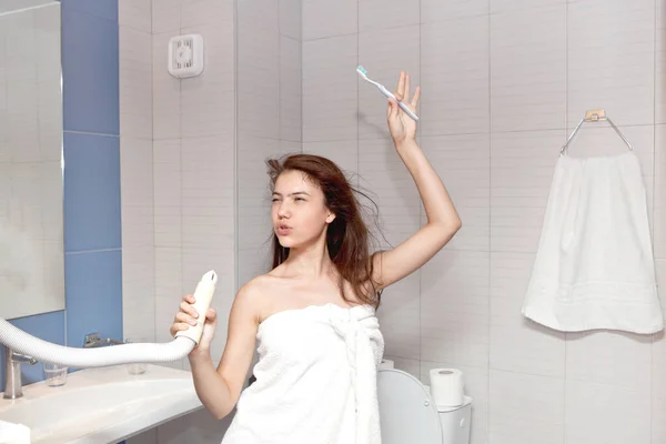 Красивая молодая девушка в белом полотенце с длинными волосами поет в ванной с феном руками. Гигиена — стоковое фото