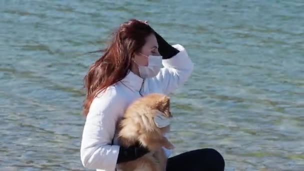 Young woman and spitz dog in a medical mask on the lake on a spring day. 4K — Stock Video