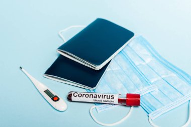 Tıbbi maskelerin, termometrenin ve pasaportların yanında mavi arka planda kan örneği ve koronavirüs içeren test tüpü.