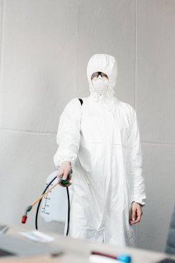 selective focus of person in white hazmat suit, respirator and goggles disinfecting workplace in office, coronavirus concept clipart