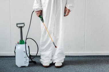 cropped view of person in white hazmat suit with disinfect in office, coronavirus concept clipart