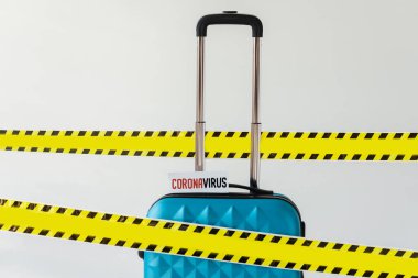 Sarı ve siyah tehlike uyarısı içeren koronavirüs kartlı mavi bavul beyaza izole edilmiş güvenlik bandı.