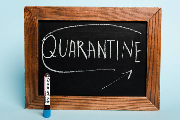 quarantine lettering written on chalkboard near test tube with coronavirus blood sample on blue background