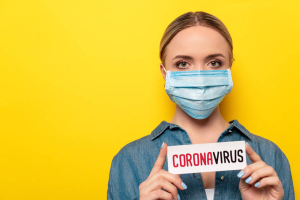 молодая женщина в медицинской маске держит карточку с коронавирусной надписью на изолированном желтом
