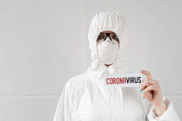 ホワイトハズマットスーツの人ですコロナウイルスの文字でカードを持っているマスクとゴーグル — ストック写真