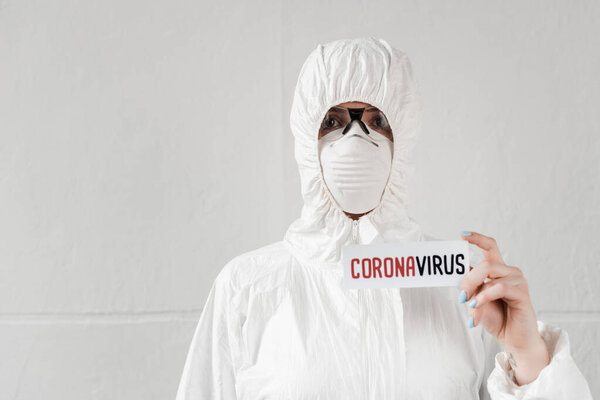 человек в белом защитном костюме, респираторе и очках, держащих карточку с коронавирусной надписью
