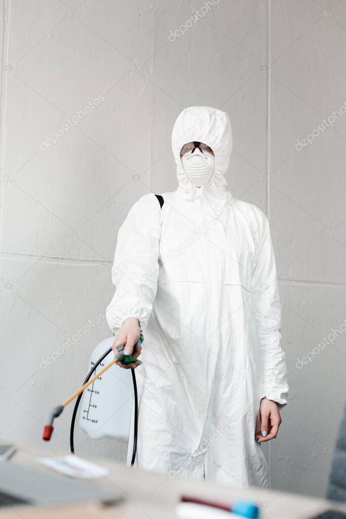 selective focus of person in white hazmat suit, respirator and goggles disinfecting workplace in office, coronavirus concept