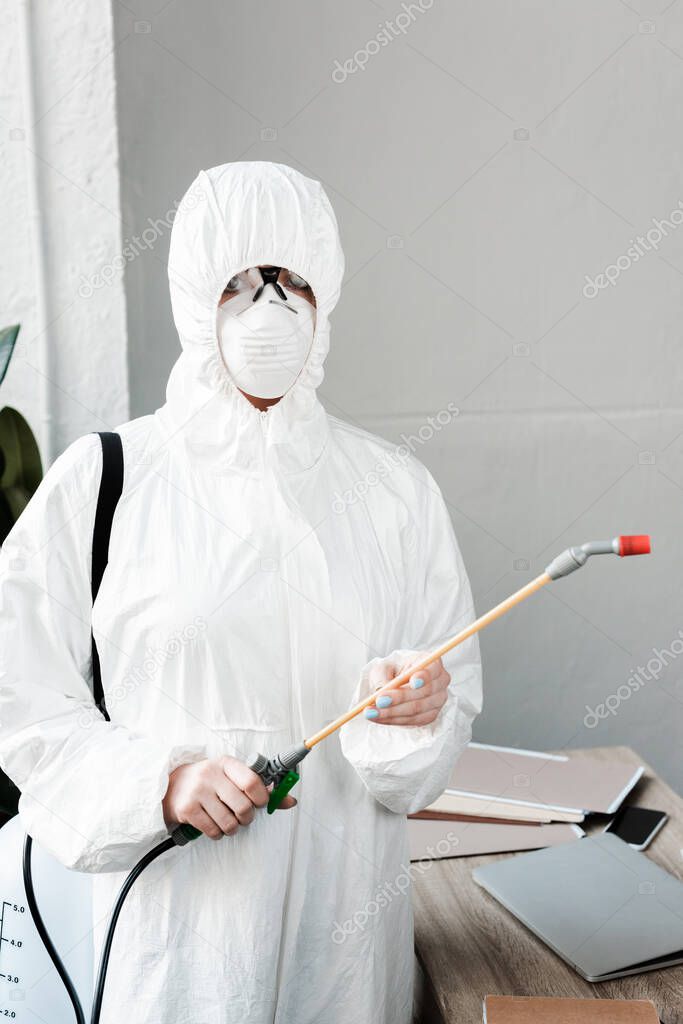 person in white hazmat suit, respirator and goggles disinfecting workplace in office, coronavirus concept
