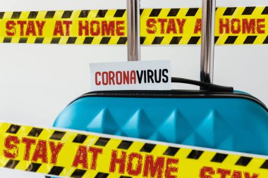 Mavi bavulu, üzerinde koronavirüs kartı olan sarı ve siyah tehlike uyarısı bandıyla kapatın ve evde kalıp beyaz resimde görünün.