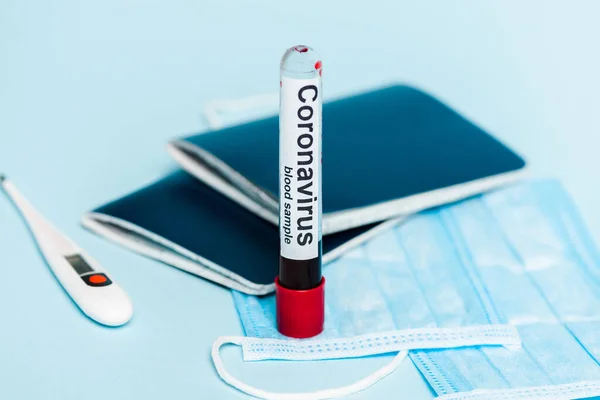 Foyer sélectif du tube à essai avec échantillon de sang et lettrage coronavirus près des masques médicaux, thermomètre et passeports sur fond bleu — Photo de stock