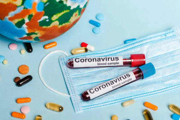 Test tubes with blood samples and coronavirus lettering near medical mask, pills and globe on blue background — Stock Photo