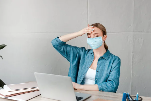 Mujer en máscara médica tocando la frente mientras trabaja con el ordenador portátil en la oficina - foto de stock