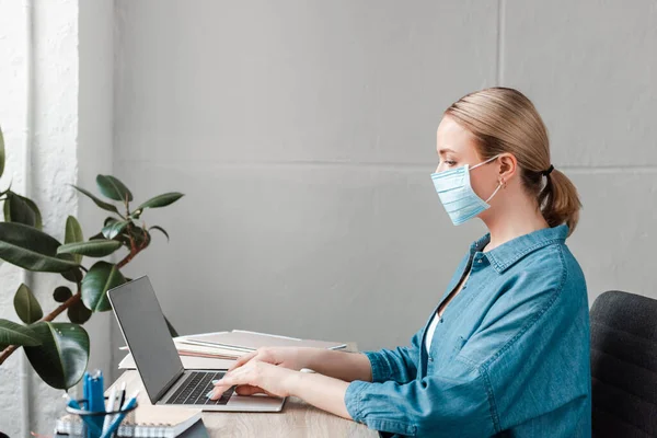 Side view of woman in medical mask working with laptop in office — Stock Photo