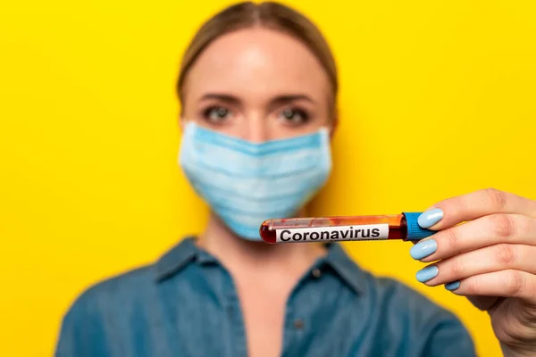 Foyer sélectif de la jeune femme dans un masque médical tenant une éprouvette avec un échantillon de sang de coronavirus sur fond jaune — Photo de stock