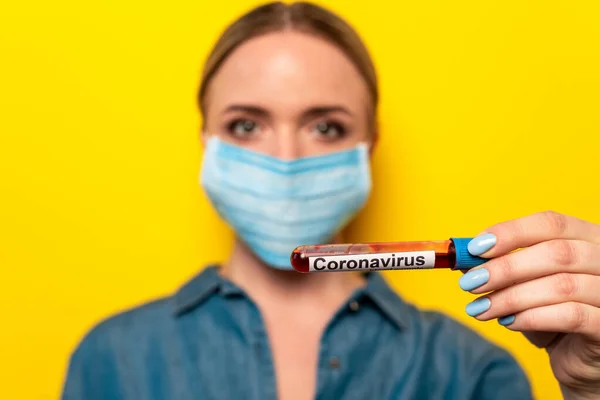 Selective focus of young woman in medical mask holding test tube with coronavirus blood sample on yellow background — Stock Photo