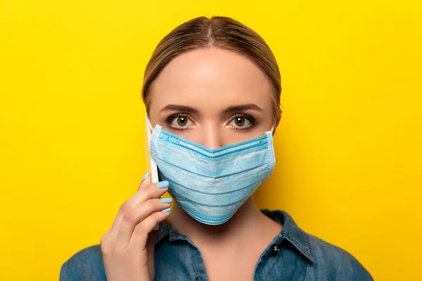 Jeune femme en masque médical parlant sur smartphone sur fond jaune — Photo de stock