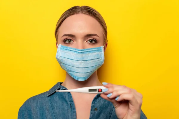 Jeune femme en masque médical tenant thermomètre sur fond jaune — Photo de stock