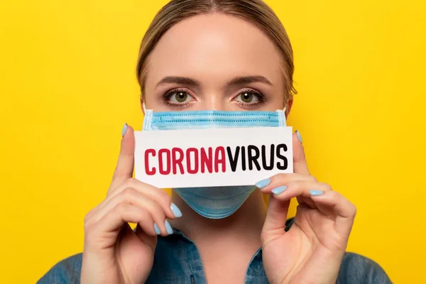 Jeune femme en masque médical tenant carte avec lettrage coronavirus sur isolé sur jaune — Photo de stock