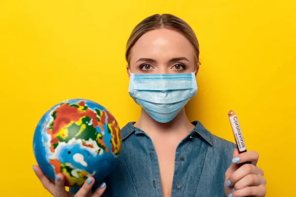 Jeune femme en masque médical tenant une éprouvette avec échantillon de sang de coronavirus et globe sur fond jaune — Photo de stock