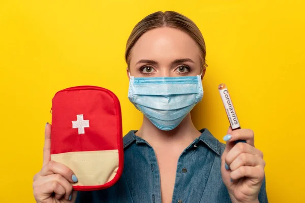 Jeune femme dans un masque médical tenant un tube à essai avec échantillon de sang de coronavirus et trousse de premiers soins sur fond jaune — Photo de stock