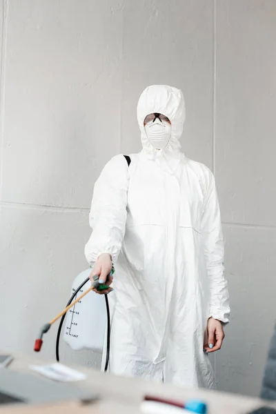 Enfoque selectivo de la persona en traje blanco hazmat, respirador y gafas desinfectando el lugar de trabajo en la oficina, concepto coronavirus - foto de stock