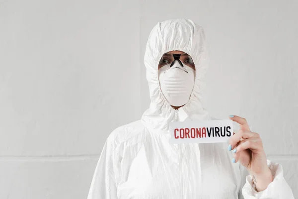 Person in white hazmat suit, respirator and goggles holding card with coronavirus lettering — Stock Photo