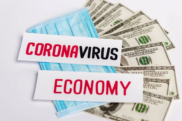Billetes en dólares, máscara médica y tarjetas coronavirus y economía sobre fondo blanco - foto de stock