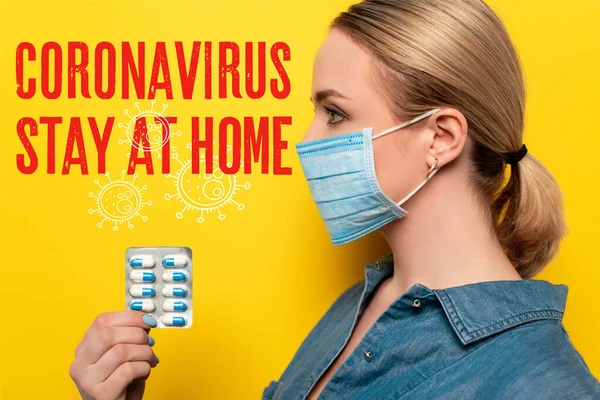 Vue latérale de la jeune femme en masque médical tenant des pilules sur fond jaune, coronavirus et rester à la maison lettrage — Photo de stock