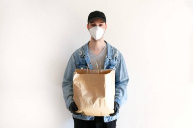 Coronavirüs salgını sırasında yiyecek ve malzeme teslimatı. Üniformalı teslimatçı. Teslimatçı bir çanta dolusu el işi kağıdı tutuyor. Koruyucu tıbbi maskeli ve lastik eldivenli adam..
