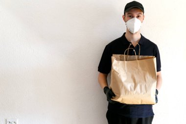 Coronavirüs salgını sırasında yiyecek ve malzeme teslimatı. Üniformalı teslimatçı. Teslimatçı bir çanta dolusu el işi kağıdı tutuyor. Koruyucu tıbbi maskeli ve lastik eldivenli adam..