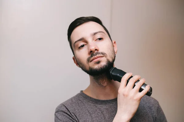 A young man with dark hair shaves his beard. A bearded man mows a beard with a trimmer.