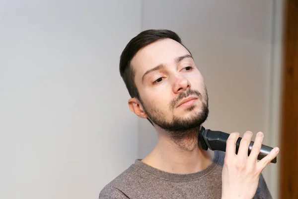 A young man with dark hair shaves his beard. A bearded man mows a beard with a trimmer.